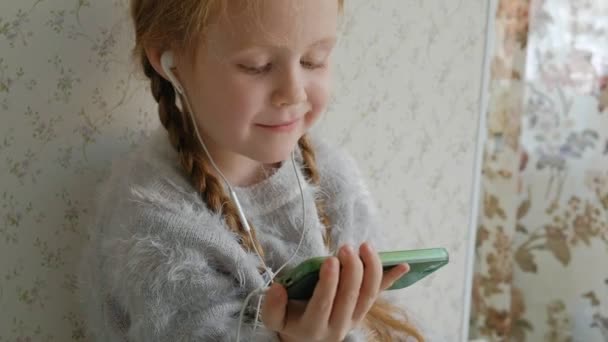 Щаслива маленька дівчинка з кісками в навушниках дивиться на телефонне відео, посміхається, сидить у кімнаті на підвіконні, покриває себе килимком, портрет — стокове відео