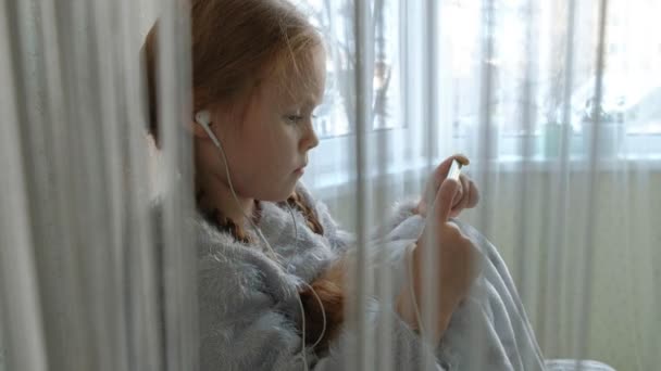 Heureuse petite fille avec des nattes dans les écouteurs regardant la vidéo du téléphone, souriant, assis dans la chambre sur le rebord de la fenêtre, se couvrant d'un tapis, portrait — Video