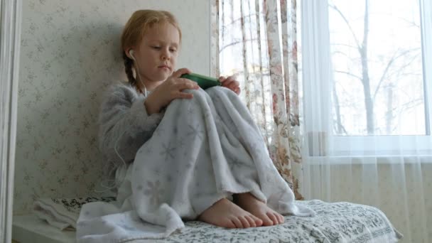 Glückliches kleines Mädchen mit Zöpfen im Kopfhörer schaut sich das Handyvideo an, sitzt in einem Raum auf der Fensterbank, versteckt eine Decke — Stockvideo