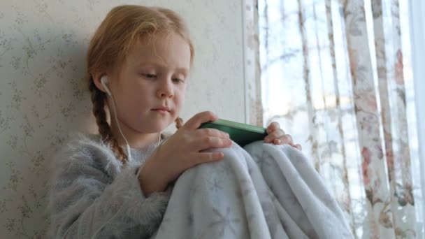 Glückliches kleines Mädchen mit Zöpfen im Kopfhörer, das sich das Handyvideo ansieht, lächelnd, im Zimmer auf der Fensterbank sitzend, sich mit einem Teppich bedeckt, Porträt — Stockvideo