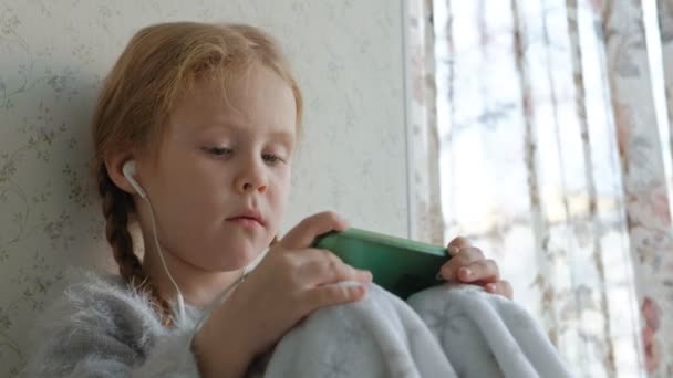 Glückliches kleines Mädchen mit Zöpfen im Kopfhörer, das sich das Handyvideo ansieht, lächelnd, im Zimmer auf der Fensterbank sitzend, sich mit einem Teppich bedeckt, Porträt — Stockvideo