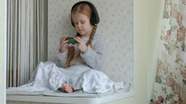 Happy little girl with pigtails in headphones looks at the phone video, sits in a room on the windowsill, hid a blanket — Stock Video