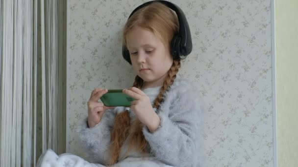 Menina feliz com tranças em fones de ouvido olhando para o vídeo do telefone, sorrindo, sentada na sala no peitoril da janela, cobrindo-se com um tapete, retrato — Vídeo de Stock