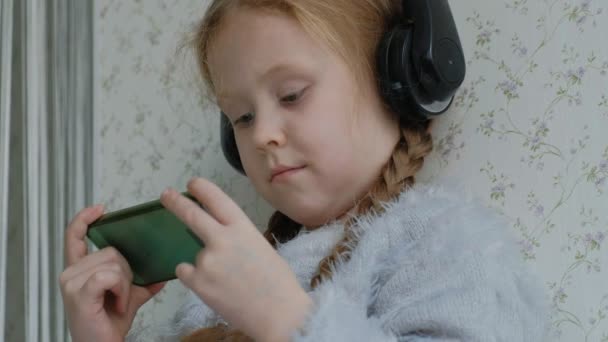 Heureuse petite fille avec des nattes dans les écouteurs regardant la vidéo du téléphone, souriant, assis dans la chambre sur le rebord de la fenêtre, se couvrant d'un tapis, portrait — Video