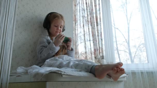 Niña feliz con coletas en los auriculares mira el video del teléfono, se sienta en una habitación en el alféizar de la ventana, escondió una manta — Vídeo de stock