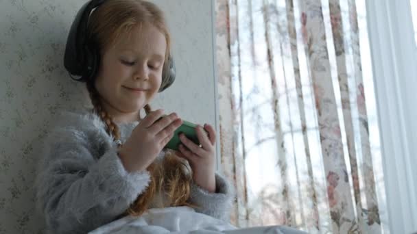 Niña feliz con coletas en los auriculares mirando el video del teléfono, sonriendo, sentada en la habitación en el alféizar de la ventana, cubriéndose con una alfombra, retrato — Vídeo de stock