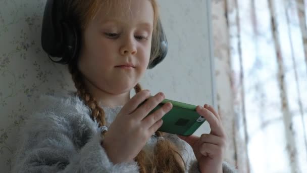 Ευτυχισμένη κοριτσάκι με κοτσίδες ακουστικά, χρησιμοποιεί το τηλέφωνο, παίζει, χαμογελαστοί, κάθεται στο δωμάτιο στο περβάζι, που καλύπτει τον εαυτό της με ένα χαλί, γκρο πλαν χέρια — Αρχείο Βίντεο