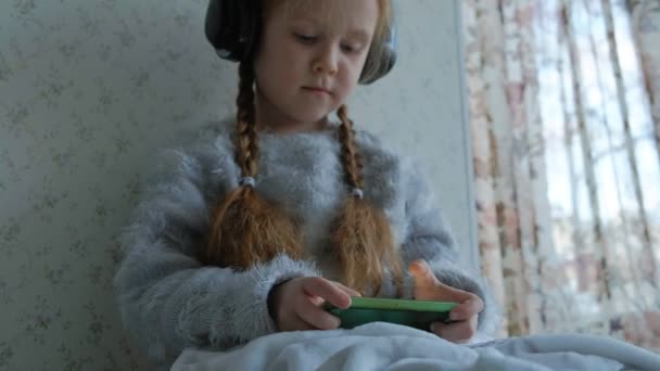 Gelukkig klein meisje met pigtails hoofdtelefoon, maakt gebruik van de telefoon, speelt, brede glimlach, zittend in de kamer op de vensterbank, die zichzelf met een tapijt, close-up handen — Stockvideo