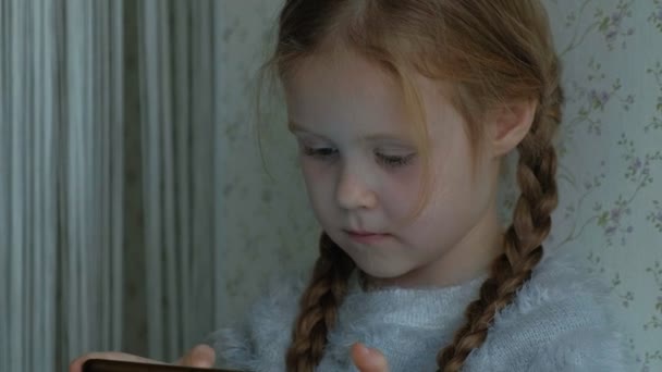 Lycklig liten flicka med flätor, använder telefonen, spelar, leende, sitter i rummet på fönsterbrädan, porträtt — Stockvideo
