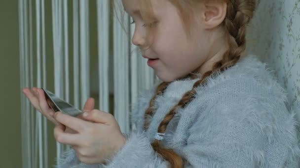 Niña feliz con coletas, utiliza el teléfono, juega, sonriendo, sentado en la habitación en el alféizar de la ventana, retrato — Vídeo de stock