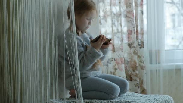 Gelukkig klein meisje met pigtails, maakt gebruik van de telefoon, speelt, brede glimlach, zittend in de kamer op de vensterbank — Stockvideo