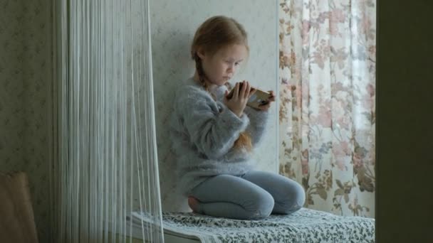 Szczęśliwy, mała dziewczynka z warkoczykami, używa telefonu, gra, uśmiechnięty, siedząc w pokoju na parapecie — Wideo stockowe