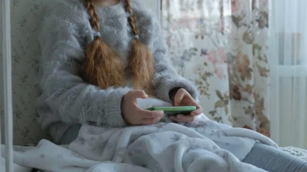 Lycklig liten flicka med flätor i hörlurar, använder telefonen, spelar, leende, sitter i rummet på fönsterbrädan, som täcker sig själv med en matta, närbild händer — Stockvideo