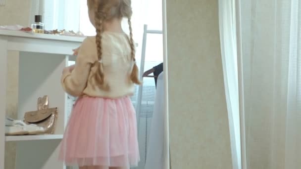 Fröhliches kleines Mädchen mit Zöpfen im rosafarbenen Rock bemalt ihre Lippen mit Lippenstift vor dem Spiegel, der Kleiderschrank ihrer Mutter — Stockvideo