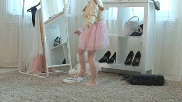 Joyeuse petite fille avec des tresses dans une jupe rose essaie sur les chaussures de mères sur les talons et danse devant un miroir avec un sac, garde-robe de mères — Video