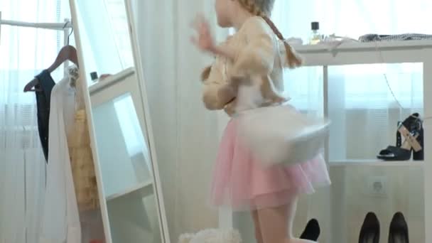 Cheerful little girl with pigtails in a pink skirt tries on mothers shoes on heels and dances in front of a mirror with a bag, mothers wardrobe — Stock Video