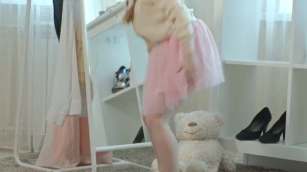 Веселая маленькая девочка с косичками в розовой юбке примеряет туфли матерей на каблуках и танцует перед зеркалом с сумкой, шкаф матерей — стоковое видео