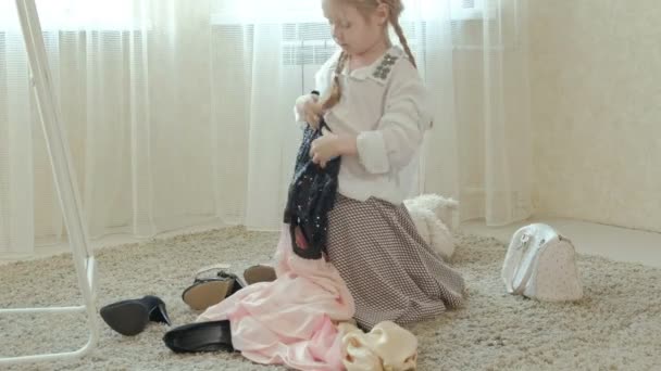 Menina alegre com tranças em uma saia rosa tenta em mães adultas roupas, vestidos, saltos altos e danças na frente de um espelho com uma bolsa, guarda-roupa mães — Vídeo de Stock