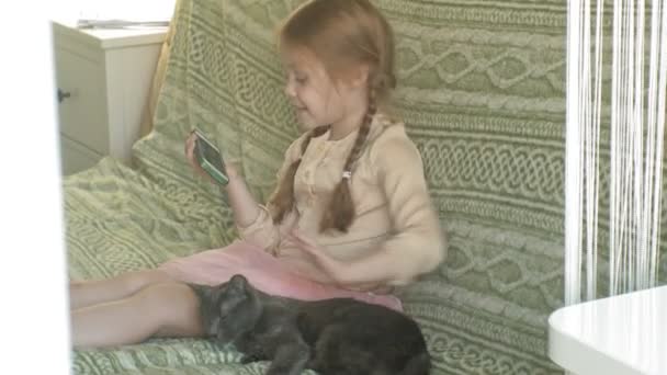 Счастливая девушка с светлыми волосами и косичками сидит на диване, используя телефон, гладит серого кота — стоковое видео