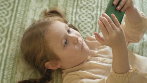 Glad tjej barn med blont hår och flätor, ligga i soffan, använder telefonen, leende, porträtt — Stockvideo