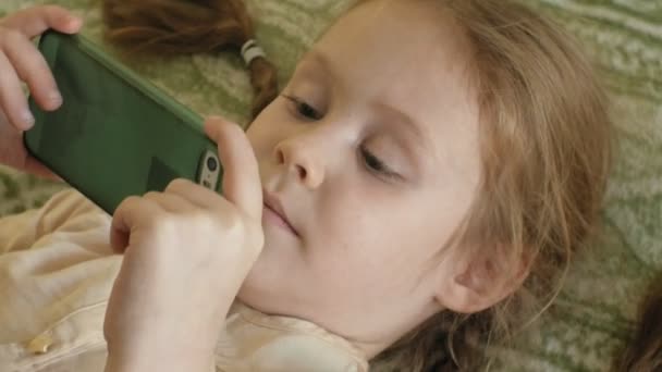 Niña feliz con el pelo rubio y coletas, acostado en el sofá, usando el teléfono, sonriendo, retrato — Vídeo de stock