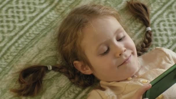 Kind van de gelukkige meisje met blond haar en pigtails, liggend op de Bank, via de telefoon, glimlachen, portret — Stockvideo