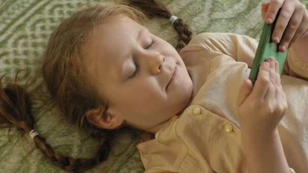 Niña feliz con el pelo rubio y coletas, acostado en el sofá, usando el teléfono, sonriendo, retrato — Vídeos de Stock