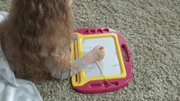 A little girl with red wavy hair lies on the floor and draws on a magnetic board. The concept of the educational process. — Stock Video