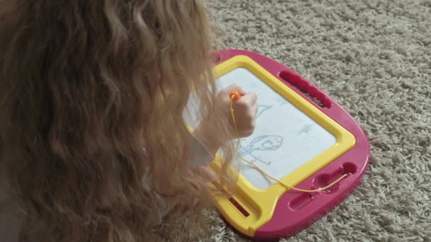 Una niña pequeña con el pelo rojo ondulado yace en el suelo y dibuja en una pizarra magnética. El concepto del proceso educativo . — Vídeo de stock