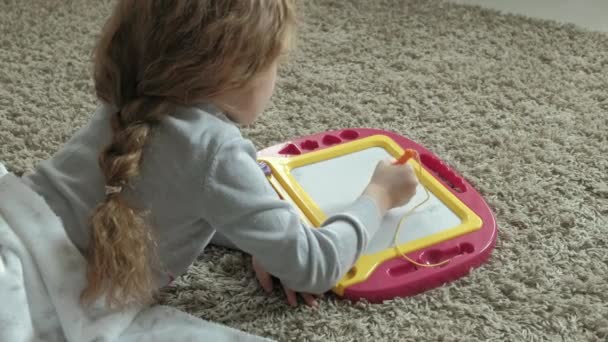 Una bambina con i capelli rossi ondulati giace sul pavimento e disegna su una lavagna magnetica. Il concetto di processo educativo . — Video Stock