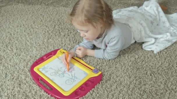 Una bambina con i capelli rossi ondulati giace sul pavimento e disegna su una lavagna magnetica. Il concetto di processo educativo . — Video Stock