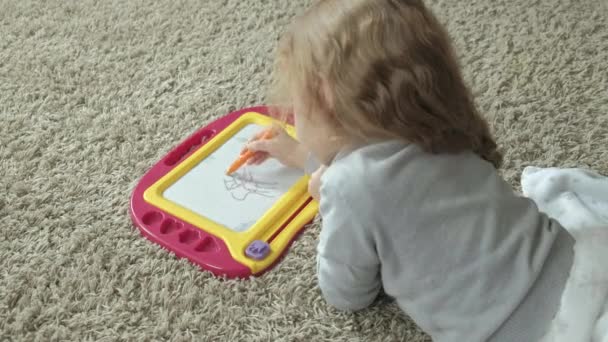 Una bambina con i capelli rossi ondulati giace sul pavimento e disegna su una lavagna magnetica. Il concetto di processo educativo . — Video Stock