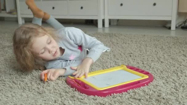 Маленькая девочка с рыжими волнистыми волосами лежит на полу и рисует на магнитной доске. Концепция учебного процесса . — стоковое видео