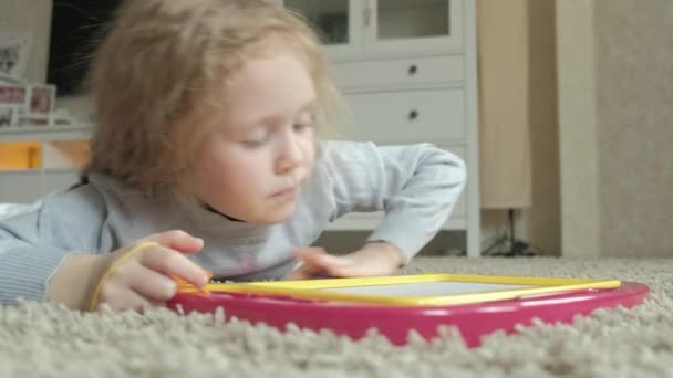 Ein kleines Mädchen mit roten welligen Haaren liegt auf dem Boden und zeichnet auf eine Magnettafel. das Konzept des Bildungsprozesses. Nahaufnahme — Stockvideo