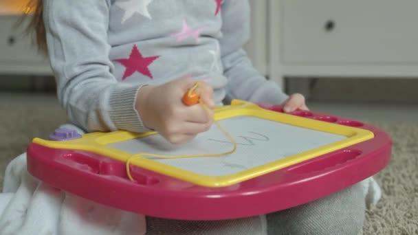 Una bambina con i capelli rossi ondulati giace sul pavimento e disegna su una lavagna magnetica. Il concetto di processo educativo. primo piano — Video Stock