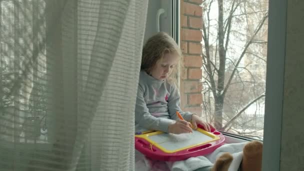 Holčička s červenou vlnité vlasy sedí na parapetu a kreslí na magnetické tabuli. Koncept vzdělávacího procesu. — Stock video