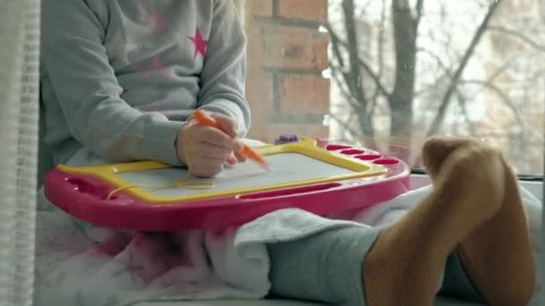 Uma menina com o cabelo ondulado vermelho senta-se no peitoril da janela e desenha em uma placa magnética. O conceito do processo educativo . — Vídeo de Stock