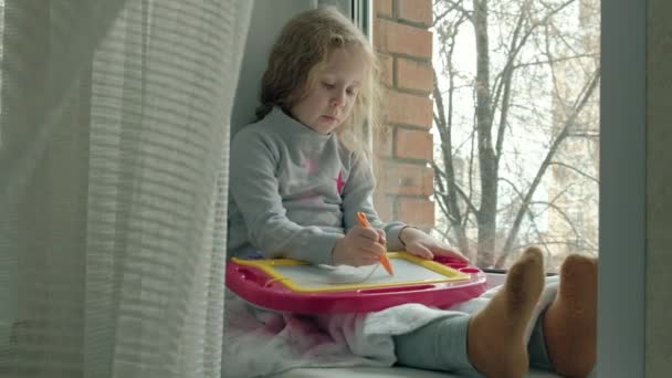 Una niña pequeña con el pelo rojo ondulado se sienta en el alféizar de la ventana y se dibuja en una placa magnética. El concepto del proceso educativo . — Vídeos de Stock