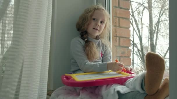 赤のウェーブのかかった髪を持つ小さな少女は窓辺に座っている、マグネット ボードに描画します。教育過程の概念. — ストック動画