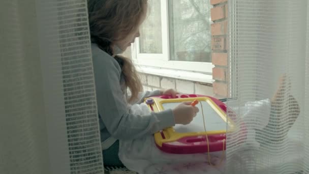 Маленька дівчинка з червоним хвилястим волоссям сидить на підвіконні і малює на магнітній дошці. Концепція освітнього процесу . — стокове відео