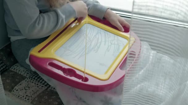 Ein kleines Mädchen mit roten welligen Haaren sitzt auf der Fensterbank und zeichnet an einer Magnettafel. das Konzept des Bildungsprozesses. — Stockvideo
