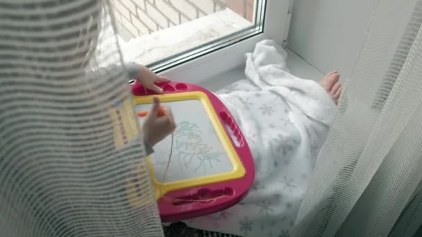 Una niña pequeña con el pelo rojo ondulado se sienta en el alféizar de la ventana y se dibuja en una placa magnética. El concepto del proceso educativo . — Vídeos de Stock