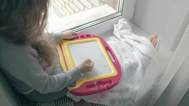 Una niña pequeña con el pelo rojo ondulado se sienta en el alféizar de la ventana y se dibuja en una placa magnética. El concepto del proceso educativo . — Vídeos de Stock