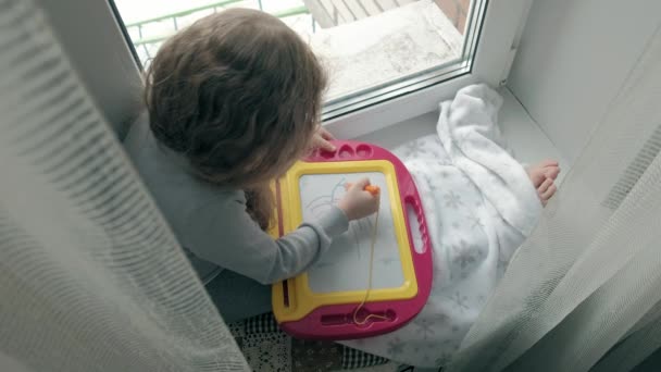 Una bambina con i capelli rossi ondulati siede sul davanzale della finestra e disegna su una lavagna magnetica. Il concetto di processo educativo . — Video Stock