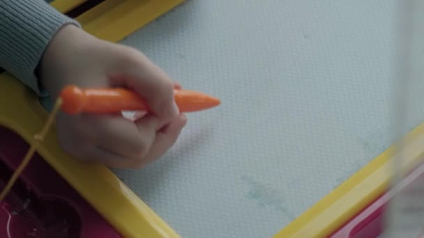 Une petite fille aux cheveux roux ondulés s'assoit sur le rebord de la fenêtre et dessine sur un tableau magnétique. Le concept du processus éducatif. gros plan — Video