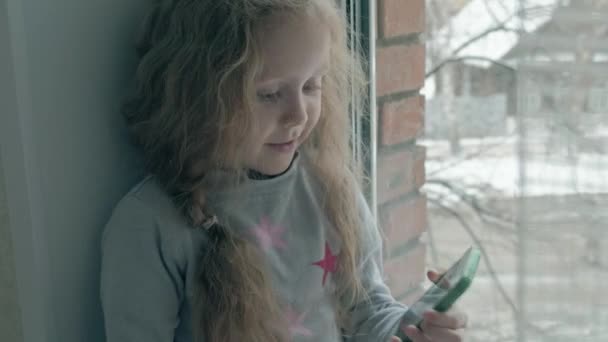 Heureuse petite fille aux cheveux roux ondulés assise sur le rebord de la fenêtre, couvrant une couverture et utilisant le téléphone, parlant, appel vidéo, portrait en gros plan — Video