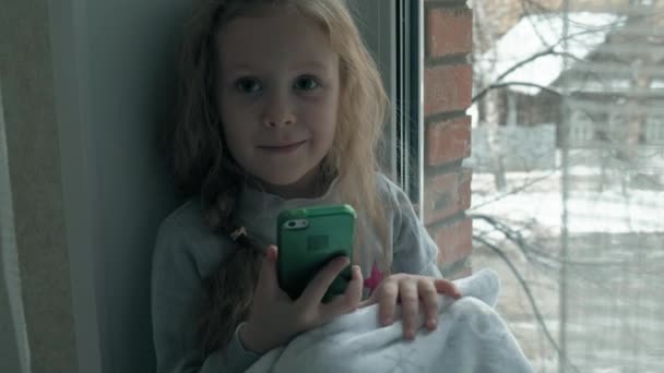 Šťastná holčička s vlnité zrzavé vlasy sedí na okenním parapetu, zahrnující deku a používání telefonu, mluvení, video volání, makro portrét — Stock video