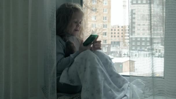 Niña feliz con el pelo rojo ondulado se sienta en el alféizar de la ventana, cubriendo una manta y usando el teléfono, hablando, videollamadas — Vídeo de stock
