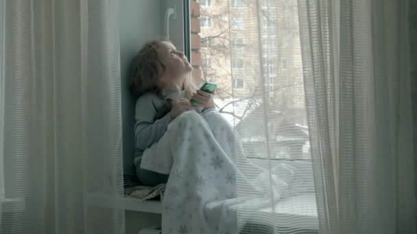 Happy little girl with wavy red hair sits on the windowsill, covering a blanket and using the phone, talking, video calling — Stock Video