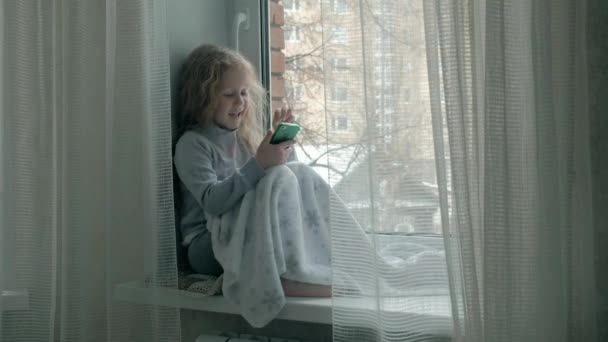 Niña feliz con el pelo rojo ondulado se sienta en el alféizar de la ventana, cubriendo una manta y usando el teléfono, hablando, videollamadas — Vídeos de Stock